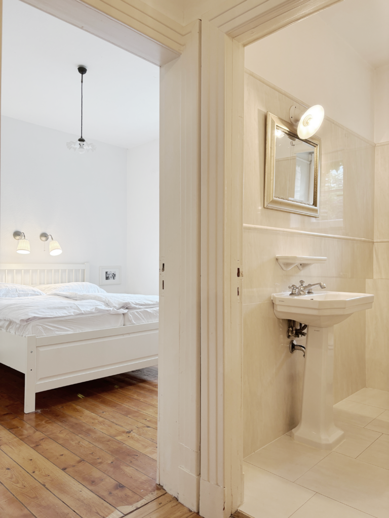 Blick vom Bad ins Schlafzimmer mit weißem Bett und Dielenboden.